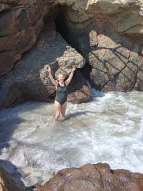 A joyful woman poses in the surf at a rocky beach, celebrating her day in the sun — a perfect moment for beach lovers and adventure seekers.