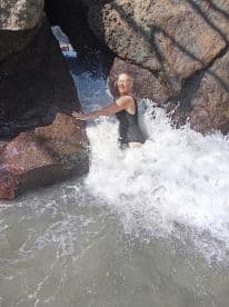 A woman joyfully interacts with ocean waves near a rocky formation, capturing the essence of adventure and fun in the water.
