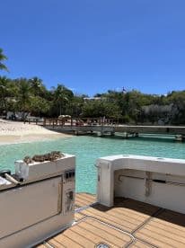 A serene view from the deck of a boat, showcasing a beautiful beach and dock area surrounded by lush greenery and clear turquoise waters.