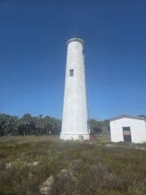 A tall lighthouse stands against a clear blue sky, surrounded by vegetation — a picturesque coastal landmark.