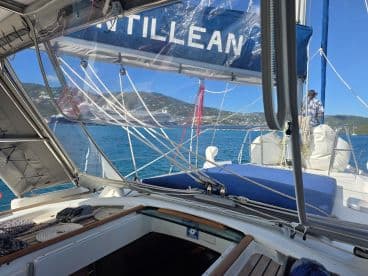 A view from the interior of a sailboat, showcasing the helm area and a crew member preparing for sailing. The serene waters and distant hills create a picturesque backdrop for a sailing adventure.