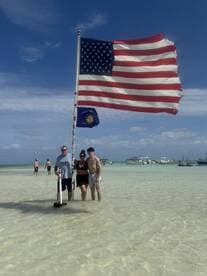 A group poses at a sandbar with an American flag, enjoying a sunny day at the beach — a perfect backdrop for memorable moments and celebrations.