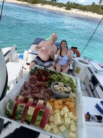 A cheerful couple enjoys refreshing cocktails and a delicious spread of fruits and meats aboard their boat, with a beautiful beach backdrop — perfect for a fun day on the water.