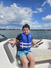A young boy enjoys a sunny day on the boat, wearing a life jacket and smiling against a backdrop of blue skies and water — perfect for family boating adventures.