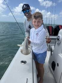 A young boy proudly holds a shark while fishing with an adult on a boat, showcasing a memorable wildlife encounter at sea.
