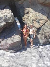 A couple poses playfully by a rock formation on a sunny beach, capturing a moment of adventure and relaxation in a tropical paradise.