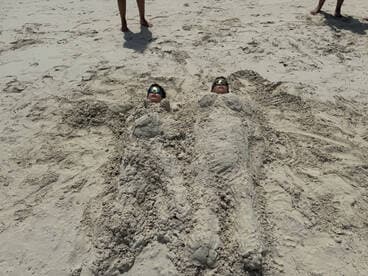 Two children enjoy a playful day at the beach, buried in sand and wearing sunglasses — a perfect snapshot of summer fun.
