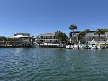 A scenic view of several boats docked along a waterfront, showcasing a beautiful day by the water with palm trees and residential buildings in the background.