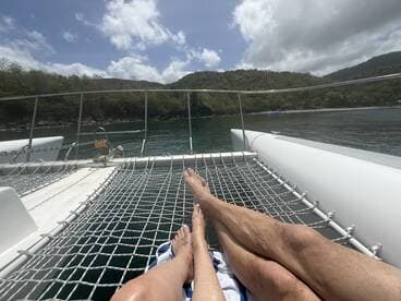Two people relax on the netting of a catamaran, enjoying the serene waters and beautiful scenery — a perfect moment for unwinding on a sailing adventure.