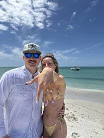 A happy couple celebrates their engagement on the beach, showcasing the ring against a picturesque backdrop of the ocean and a boat in the distance.