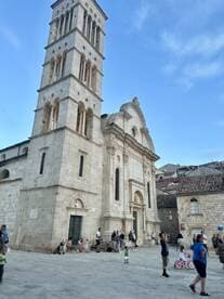 A lively scene featuring a historic building with various people, including children, enjoying the area. The architecture and atmosphere suggest a vibrant public space.