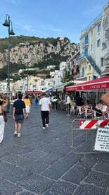 A bustling street scene with people walking past various bars and restaurants, set against a backdrop of rocky hills. The atmosphere captures the lively essence of a popular tourist destination.