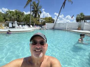 A man takes a selfie in a pool surrounded by other guests enjoying the water on a sunny day.