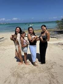 A group of four women and a girl enjoy a playful moment on the beach, holding adorable piglets — a memorable wildlife encounter in a tropical paradise.