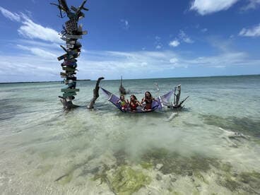 A group of friends relaxes in a hammock at a picturesque beach, surrounded by clear waters and a unique signpost — a perfect spot for fun and relaxation.