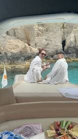 A couple enjoys a moment of celebration on a boat, wrapped in towels and sharing snacks against a stunning coastal backdrop — perfect for romantic getaways or special occasions on the water.