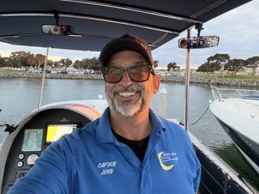 Captain John smiles from the helm of his boat, showcasing the control console and a scenic marina backdrop — perfect for boating adventures.
