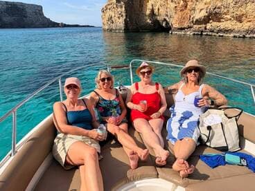 A cheerful group of friends enjoys cocktails and sunbathing on a boat, surrounded by stunning turquoise waters and rocky cliffs — perfect for a relaxing day on the water.