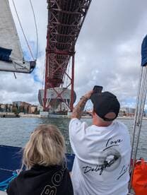 A couple enjoys a sailing trip under a stunning bridge, capturing the moment with a smartphone — a perfect blend of adventure and sightseeing.