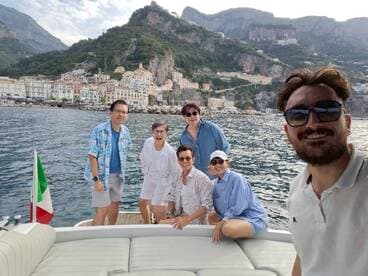 A cheerful group poses for a selfie on a boat with a stunning coastal backdrop, capturing the joy of their boating adventure.