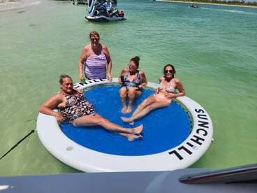 A lively group of friends enjoys a sunny day on a floating mat in clear waters, perfect for relaxation and fun in the sun.