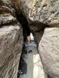 A person explores a rocky passageway leading to a sandy beach, showcasing the beauty of natural landscapes and adventure in coastal areas.