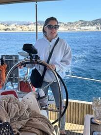 A woman stands at the helm of a sailboat, navigating through beautiful waters with a scenic backdrop — perfect for sailing adventures.