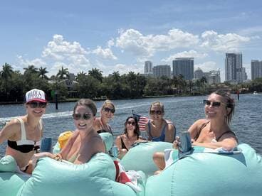 A lively group of friends enjoys a fun day on the water, relaxing on floating loungers with cocktails in hand, set against a stunning city skyline.