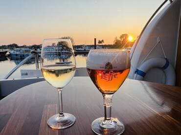 Two glasses of wine and beer sit on a table aboard a boat, with a stunning sunset reflecting on the water — perfect for a relaxing evening on the water.