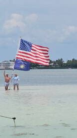 A couple proudly displays the American and Conch Republic flags while standing in shallow water, capturing a festive moment in a beautiful beach setting.