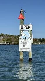 A sign for Cabbage Key, indicating it's open for business, with a scenic waterfront backdrop. Ideal for visitors seeking dining and relaxation options in a picturesque setting.