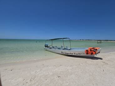 A small tour boat named 'Rio Lagartos Tours' is anchored on a beautiful beach, with guests enjoying the clear waters nearby — perfect for a day of exploration and adventure.
