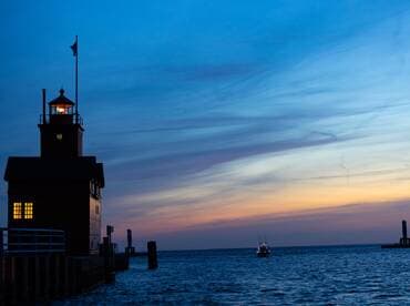 A picturesque sunset view featuring a lighthouse and a boat approaching the harbor, creating a serene maritime atmosphere.