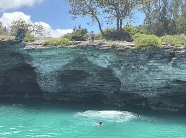 A group of people enjoys a sunny day by the cliffs, with one person swimming in the stunning turquoise waters — a perfect spot for adventure and exploration.