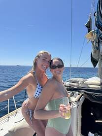Two friends enjoy a sunny day on a sailboat, celebrating with drinks and smiles against a stunning ocean backdrop.