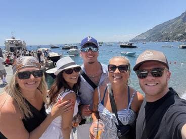 A cheerful group of friends poses for a selfie by the waterfront, surrounded by boats and stunning coastal scenery — perfect for memorable summer adventures.