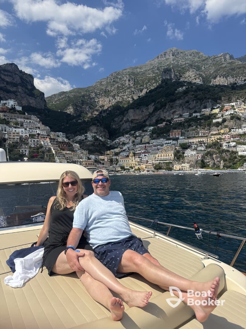 A couple relaxes on the deck of a boat, enjoying the stunning views of Positano's colorful cliffside homes under a bright blue sky — a perfect moment for a romantic getaway.