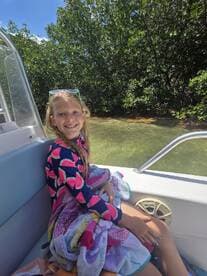 A young girl smiles while sitting on a boat surrounded by lush mangroves, showcasing a fun day on the water with towels in hand.