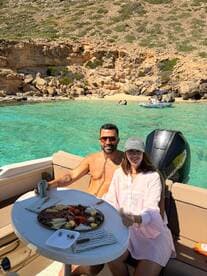 A couple enjoys a delightful spread of snacks on a boat, surrounded by stunning turquoise waters and rocky cliffs — perfect for a relaxing day on the water.