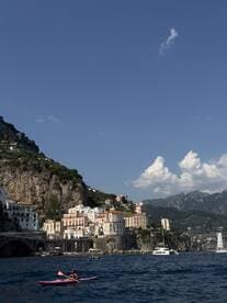 A kayaker paddles along the stunning Amalfi Coast, with colorful cliffside villages and a picturesque backdrop of mountains and blue skies.