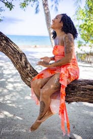 A joyful woman sits on a tree branch by the beach, enjoying the serene surroundings. The image captures a moment of happiness in a tropical setting.