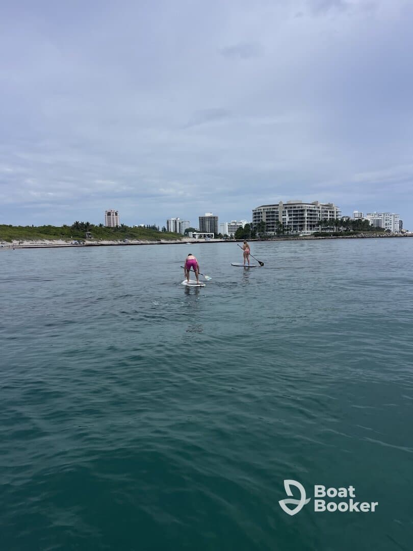 Two individuals enjoy paddleboarding in calm waters, with a scenic coastline and buildings in the background — a perfect day for water sports.