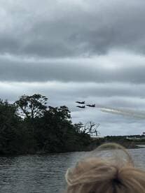 A stunning view of an airshow featuring fighter jets flying in formation over a scenic waterway, with a glimpse of a spectator enjoying the spectacle from a boat.