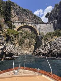 A stunning view of a boat navigating through a narrow passageway beneath a historic bridge, surrounded by rugged cliffs and lush greenery — a picturesque moment on the water.