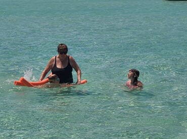 A family enjoys a fun day in the water, with one adult assisting a child on a float while another child swims nearby — perfect for memorable summer adventures.