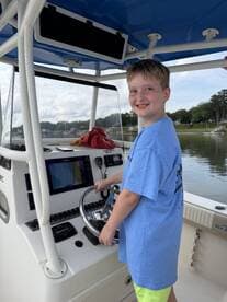 A young boy smiles while steering the helm of a boat, showcasing the excitement of learning to navigate on the water.