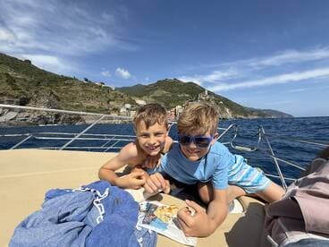 Two boys enjoy a snack on the deck of a boat, with a picturesque coastline in the background — a perfect moment of fun on the water.