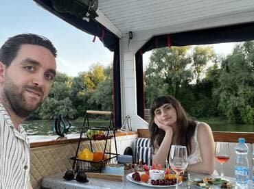 A couple enjoys a romantic dining experience on a boat, complete with cocktails and a beautiful riverside view — perfect for intimate getaways.