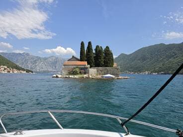 A picturesque view of a small island with lush trees and a historic building, set against a backdrop of stunning mountains and clear blue skies — a perfect destination for boat tours.