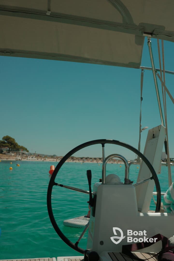 A close-up view of the helm and console of a boat, showcasing the steering wheel against a backdrop of clear turquoise waters and a sandy beach.